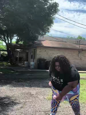A woman in colorful leggings and a black t-shirt with text is lifting pink dumbbells outside in front of a building.