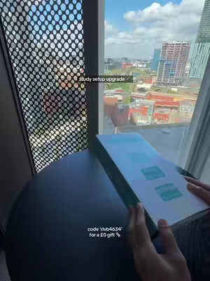 The cover image shows a person's hands holding a rectangular white box with blue and turquoise accents in front of a window. The background outside the window depicts a cityscape with tall buildings and a partly cloudy sky. The setting is a room with a dark, round table appearing in the foreground. Text overlays are present.