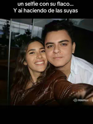 A selfie featuring a smiling couple, the woman on the left is wearing a brown jacket and the man on the right is wearing a white shirt. The image is taken in a low-light setting.