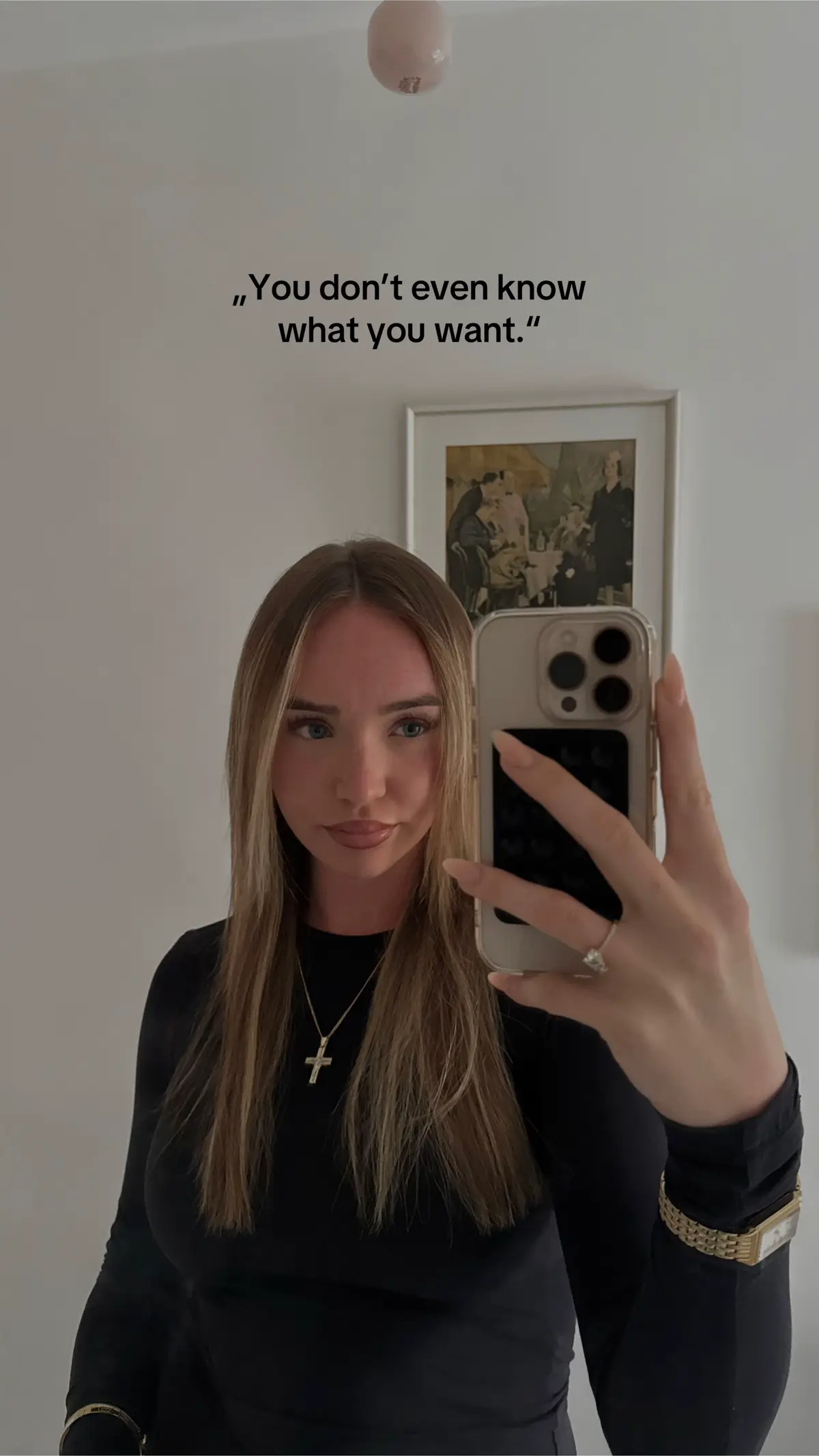 A woman takes a selfie in what appears to be a room. She is holding a phone with her hand up to take the photo. There's a text overlay that says "You don't even know what you want." She is wearing a black long sleeve top with a necklace. In the background there is a picture on the wall.