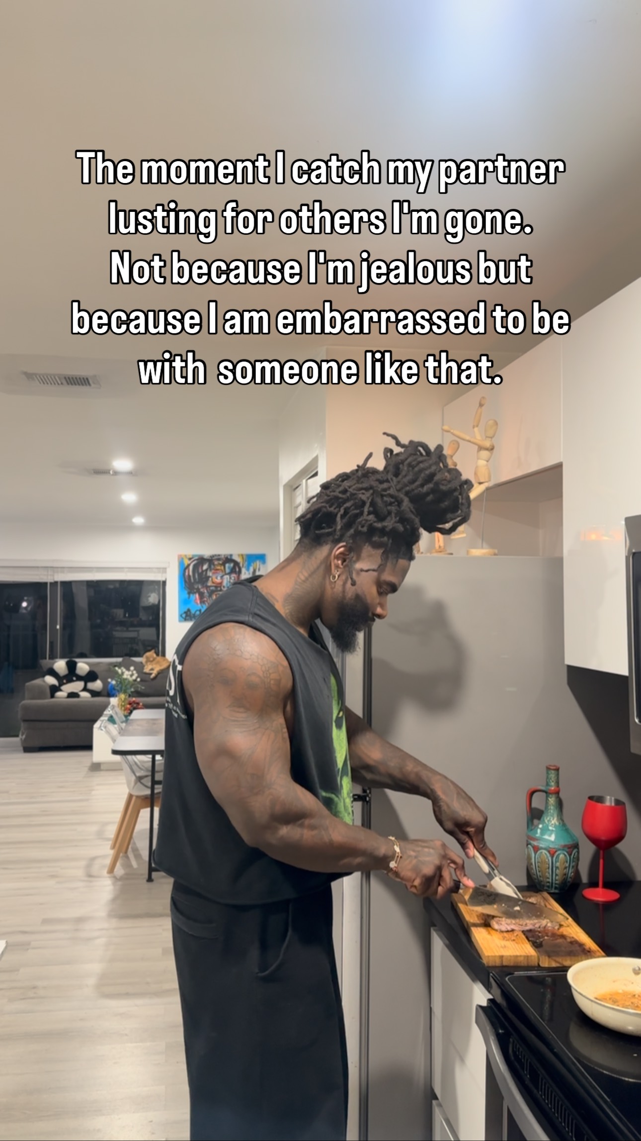 A man is shown cooking in a kitchen. He has tattoos on his arm, and he's holding a knife in his right hand while preparing food on a small wooden board. Behind him, there's a glimpse of living room decor. The image is taken in a well-lit interior setting.