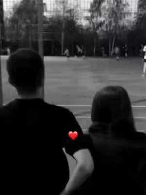 A black and white image of a couple from behind, with the man in the foreground and the woman slightly behind on the right. A red heart emoji is on the man's sleeve.