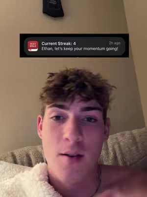 A young man is centered in the frame, with a soft, neutral-toned background. He has short, tousled brown hair and is looking directly at the camera with a neutral expression. He appears to be sitting, possibly on a couch, and is wearing a white, fluffy robe or a blanket. A notification from an app or social media is displayed above his head, featuring the Holy Bible app's logo. The notification includes the text 'Current Streak: 4' and 'Ethan, let's keep your momentum going!'
