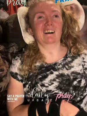 A woman with blonde curly hair sits in what appears to be a chair or medical setting, smiling with her mouth open. Text overlays read 'daily update' and 'Say A Prayer With Me'.
