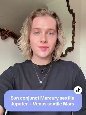 A selfie-style video shows a person with long, wavy blonde hair wearing a black t-shirt and two necklaces. The person is smiling and facing the camera, with a light-purple and white box at the bottom that contains some text and the TikTok logo. The background includes a partially visible ornate mirror and a white wall.
