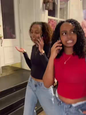 Two young women, both with dark hair, stand outside a building, talking on cell phones. One wears a red top while the other is wearing a black top. They're in mid-conversation, gesturing with their hands.