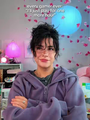 A young woman is centered in the frame with arms crossed. She's wearing an oversized purple hoodie, a silver necklace, and a ring on her finger. Behind her is a wall decorated with paper flowers, a blue orb light, and a pink lamp on a shelf. The expression suggests a playful lie.