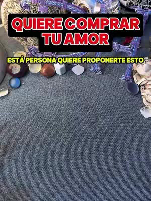 The image features a close-up of various objects and text overlaid. The objects include small, colorful stones and decorative items. Above these, a black banner displays the text 'QUIERE COMPRAR TU AMOR' in red. Below, another line of text in yellow reads 'ESTÁ PERSONA QUIERE PROPONERTE ESTO.' The background is a textured gray surface.