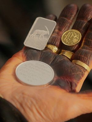 Close-up of a hand holding various precious items: a silver rectangular ingot with a gazelle engraving, a gold coin, and gold rings. The hand is painted with henna, displaying detailed patterns and dark coloration.