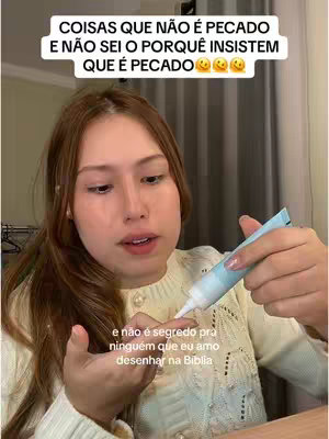 A woman with long brown hair looks directly at the camera, holding a tube of cream and applying it to her finger. Text overlays are at the top and the bottom of the image. The background is a room setting with curtains and a glimpse of a clothing rack.