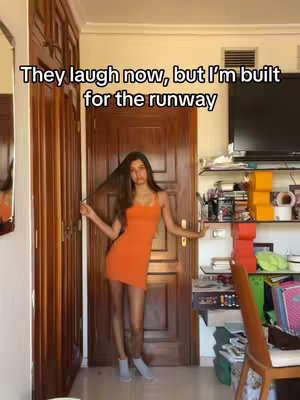 The image shows a young woman in a fitted orange mini dress standing in what appears to be a doorway. The background includes a wooden door, a closet, and elements of a room, such as shelves with items and a television.