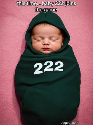 A baby wrapped in a green hoodie-like covering with the number "222" printed on it. The baby is sleeping with eyes closed, resting on a pink blanket. There is text at the top that reads "this time... baby 222 joins the game".
