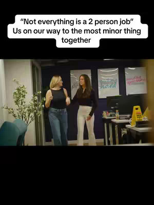 Two women are walking together in a brightly lit office space, holding hands, and smiling, indicating friendship or collaboration. The text overlay at the top reads, "'Not everything is a 2 person job' Us on our way to the most minor thing together."