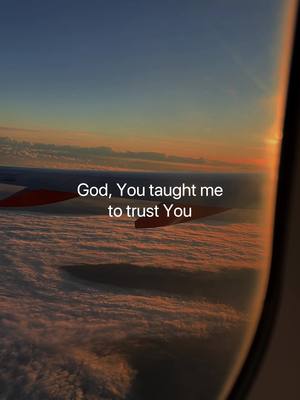 An aerial view from a plane window shows the wing of the aircraft. Below, a sea of clouds is illuminated by the setting sun, creating a warm, golden hue. The window's frame and the wing's detail are visible, with text overlayed in the top-center: 'God, You taught me to trust You'.