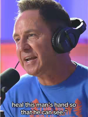 A man with short hair and a microphone. The image appears to be a screenshot from a video or podcast, as indicated by the headphones and microphone in front of the man. He's wearing a blue t-shirt and has an animated expression. The background is a blended purplish color.