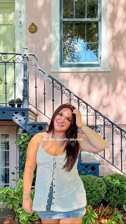 A young woman with long, dark hair poses outdoors, smiling, in front of a pink building with a green door. The background features stairs, foliage, and a window reflecting greenery. She wears a white top and denim skirt. The hook text is overlaid at the bottom.