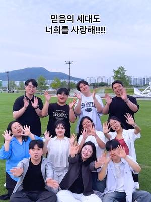 A group of young adults are in a park-like setting with green grass and trees in the background. They are waving at the camera. Text overlayed on the top of the image in Korean.