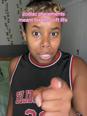 A close-up shot of a person with short, styled hair and a red and black basketball jersey, pointing directly at the camera. Text overlaid on the image reads 'zodiac placements meant for the soft life'. The background features a bed and trailing vines.