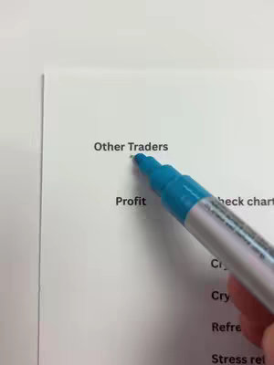 A close-up shot features a white sheet of paper with the words 'Other Traders' and 'Profit' visible. A turquoise marker pen is being used to underline the text 'Other Traders'. The background is a plain white surface, providing a clean and focused composition.