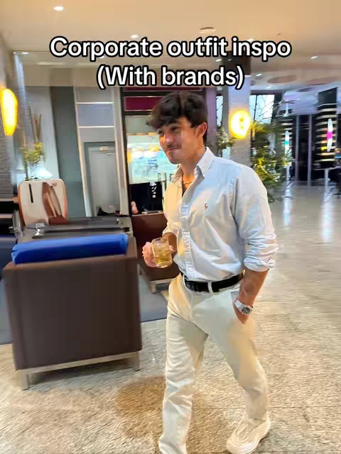 A man in a light blue button-down shirt and cream-colored pants stands in what appears to be a hotel lobby or similar setting. He holds a glass of amber liquid and smiles. The background includes an interior with seating and a window.