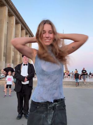 A woman with long blonde hair stands in the foreground with her arms raised behind her head, smiling towards the camera. In the midground, a man in a tuxedo stands with a stoic expression. Buildings and people are visible in the background, with the Eiffel Tower partially visible.