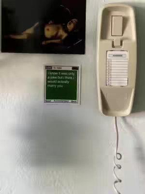 The image shows a wall with three items: a framed picture of a nude person, a small square with text, and a cream-colored wall phone. The picture depicts a person lying down, with some body exposed. The square has white text against a dark green background, and there is a phone hanging below.