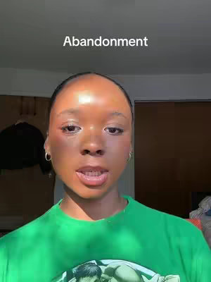 A close-up shot of a person's face looking directly at the camera, with the word "Abandonment" overlaid at the top. The person has dark skin, styled hair, and is wearing a green shirt. The background includes shadows and interior elements like a closet door. The lighting is slightly harsh, with direct sunlight on the person's face.