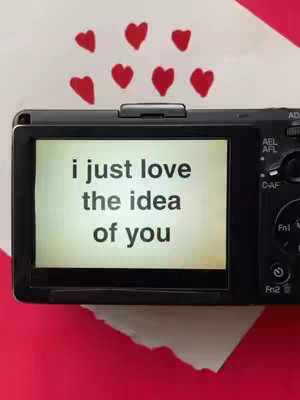 The image features a digital camera displaying the message 'i just love the idea of you' in a handwritten-style font. Above the camera, red heart doodles are scattered across a white background. The camera and message overlay the flat red surface of an extended piece of paper, positioned on a surface with a torn edge.