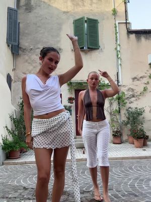 Two young women strike poses in an outdoor setting. One wears a white top and patterned skirt, raising her arm, and the other wears a halter top and white pants, posing beside her. The scene takes place in a courtyard with stone buildings and greenery. The lighting is natural.
