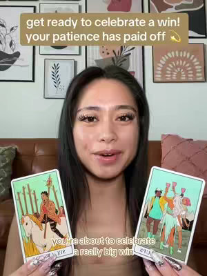 A woman with long dark hair is centered in the frame, facing the camera. She is holding two tarot cards, one in each hand, and is smiling at the viewer. Over her head is a text overlay with a gold background.