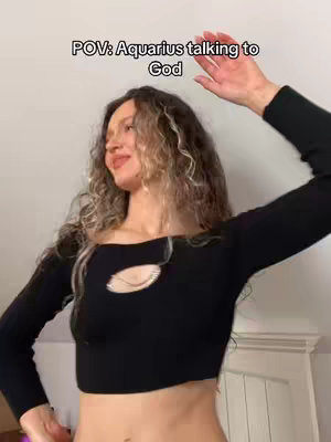A woman with curly hair and a black crop top is the central focus, posed with her arm raised as if gesturing towards the upper-right. The background is minimalist with a neutral wall and a glimpse of white furniture.
