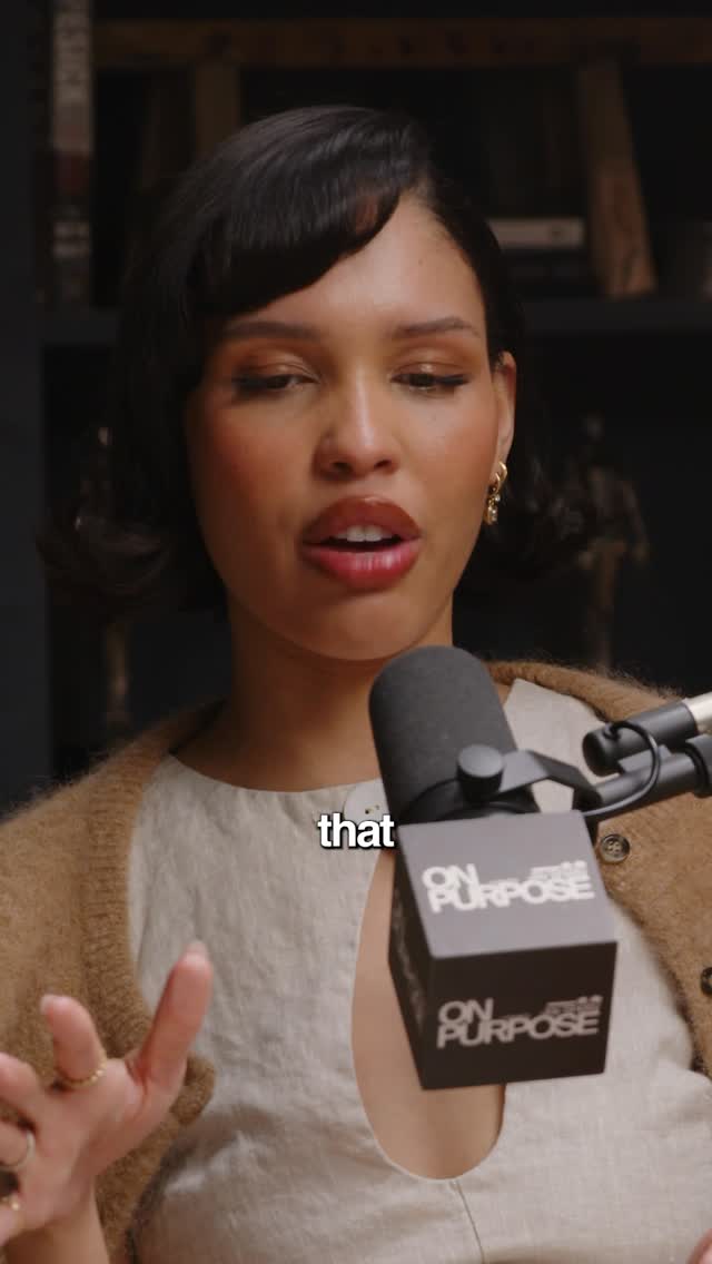 A medium shot shows a woman with dark hair and open mouth speaking into a microphone. She has earrings and a sweater. The background is blurred, showing a shelf.