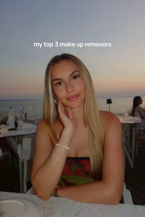 A selfie of a woman with long blonde hair holding her hand near her face, looking at the camera. She is at a lit restaurant on the water's edge at dusk. The sky is a gradient of orange and blue.