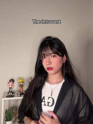 A young woman with long black hair, wearing a white t-shirt and a black sheer jacket, stands in front of a beige wall. The text "The Introvert" is displayed above her head. Behind her are small figurines on a shelf.