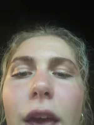 A close-up shot captures the face of a young woman with blonde hair. Her eyes look downwards, and her lips are slightly parted. The background is dark.