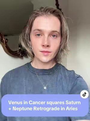 A selfie-style image featuring a fair-skinned male with long, light-brown hair, looking directly at the viewer. He's wearing a blue-gray t-shirt and a small silver necklace, the backdrop is a light-colored wall and a decorative ornate mirror frame. A text overlay at the bottom of the image reads "Venus in Cancer squares Saturn + Neptune Retrograde in Aries" within a rounded rectangle with a blurred background.