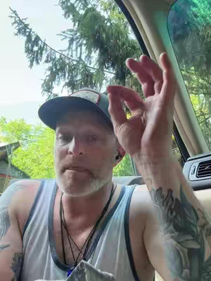 A selfie-style shot taken from inside a car, with the man's face in focus. He is wearing a baseball cap and a tank top. He is making an "okay" sign with his right hand. Behind him there’s a peek of greenery, suggesting a natural outdoor setting. The lighting appears to be natural.