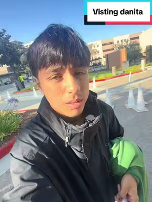 A selfie-style shot of a young man standing outside a building. He is facing the camera directly. The background includes a modern building, trees, and cones set up for traffic. The sky is clear, the lighting daylight. The man is dressed in dark clothes with a green item in his hand and a black jacket on. The video title is in a rectangle in the top right corner