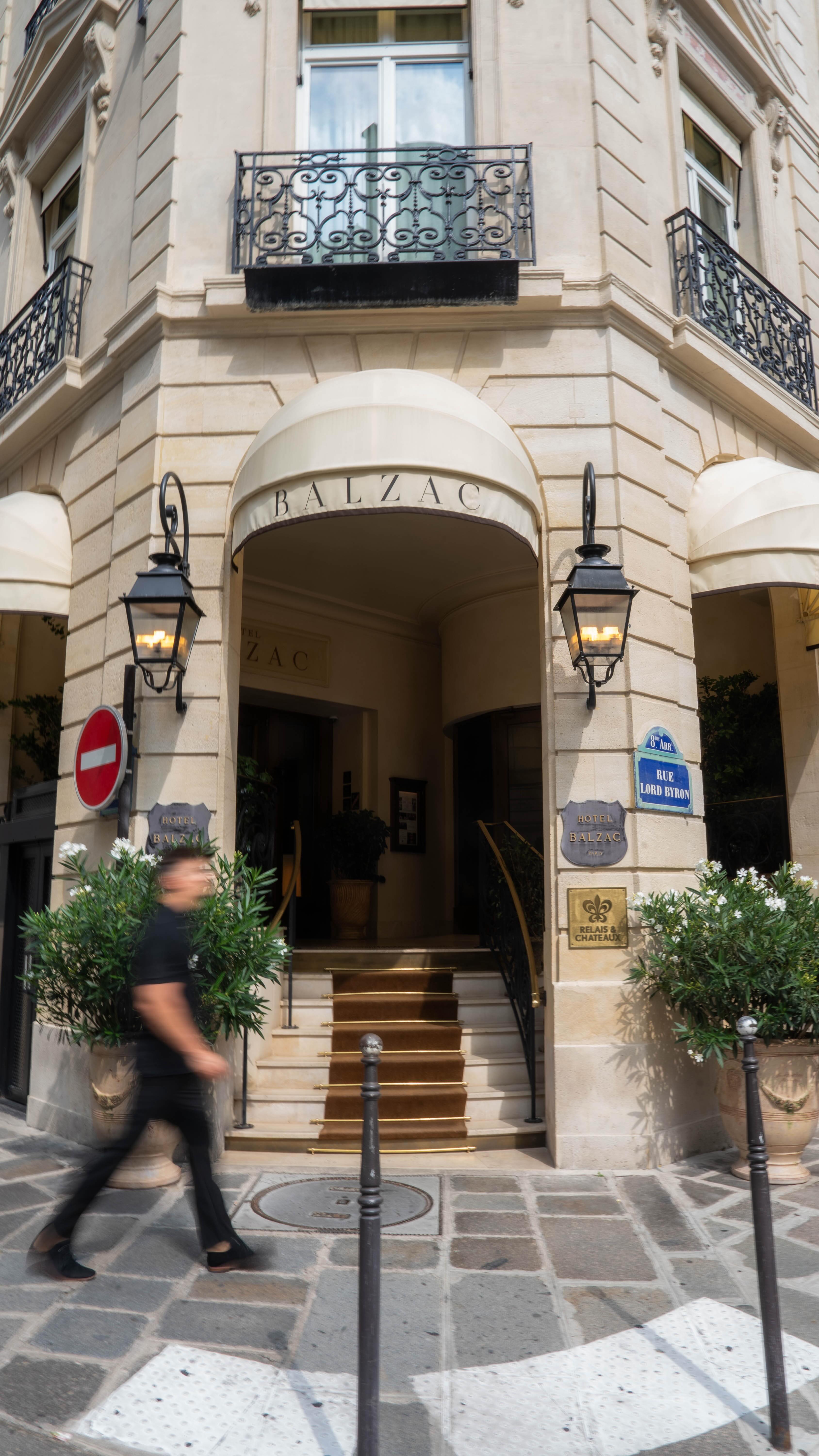 View of the entrance to the Hotel Balzac in Paris. The building is made of light-colored stone with ornate architectural details. An awning at the entrance displays the hotel name. A person is walking by in front of the entrance, they're blurred due to motion.