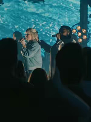 The cover image shows a live performance with three people on stage, with two women in the center, each holding a microphone and singing. They are illuminated by stage lighting. A projection of a water-like pattern serves as the background behind the performers. The audience is visible in the foreground as silhouettes.