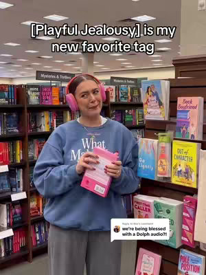A woman stands in front of shelves filled with books in a bookstore, wearing pink headphones and a blue sweatshirt. She holds a pink book and looks at the camera with a playful expression. Text saying '[Playful Jealousy] is my new favorite tag' is overlaid at the top.