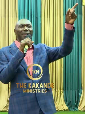 A man in a blue suit jacket and pink shirt is speaking into a microphone, with an arm raised and index finger pointing upwards. The background is a colorful mix of yellow, teal and green curtains, and there's a circular logo with text in the image "THE KAKANDE MINISTRIES".