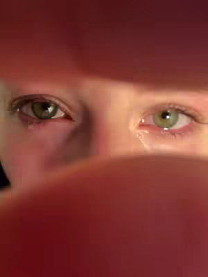A close-up shot of eyes, framed by red tones, which look like fingers. The eyes are green, with visible veins and a hint of tears in the lower lids.