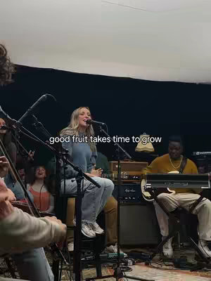 A live music performance cover image featuring a female vocalist in a denim shirt and jeans sitting on a stool, singing into a microphone. Surrounding her are other band members playing various instruments. The lighting is dim, with a dark background and visible music equipment. The text overlay reads 'good fruit takes time to grow'.
