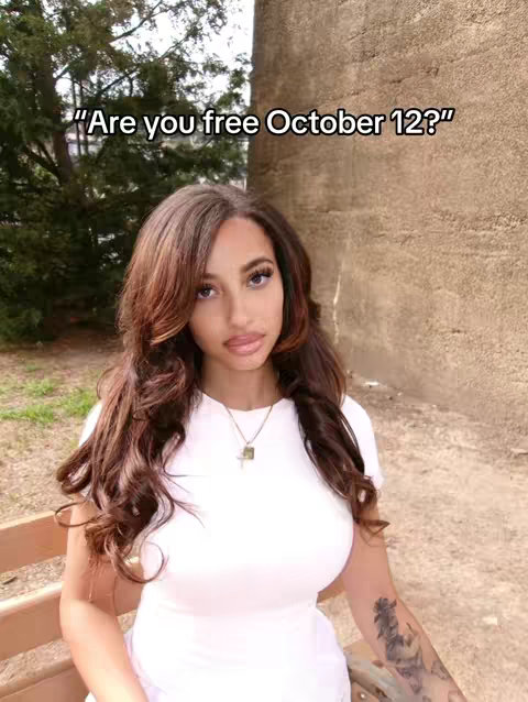 A woman with long, wavy brown hair and a white top sits on a wooden bench in front of a neutral-toned background. Overlaid text asks, "Are you free October 12?"