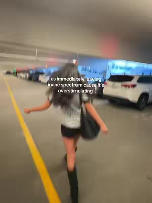 A person with long hair is running toward the camera out of a parking structure. Text overlaid on the image says they are leaving Irvine Spectrum because it is overstimulating. Cars are parked along the sides with a yellow stripe on the lane.