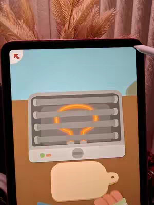 An iPad screen displays a cooking game interface. The game features a cartoon-style design, with a light blue background and a cartoon grill in the center emitting a yellow light. Below the grill is a wooden cutting board. The overall composition is simple, using a limited color palette.