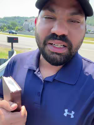 A man with a beard wearing a dark Under Armour polo shirt and a black baseball cap stands outdoors, holding a small book. Behind him is a blurred background with trees and a building. The man is looking at the camera. He is holding a book.