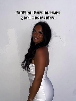 A woman with long dark hair and a tan stands in front of a plain, off-white wall. She is wearing a white dress and looking slightly to the side with a subtle smile. The text overlayed at the top of the image says, "don't go there because you'll never return".
