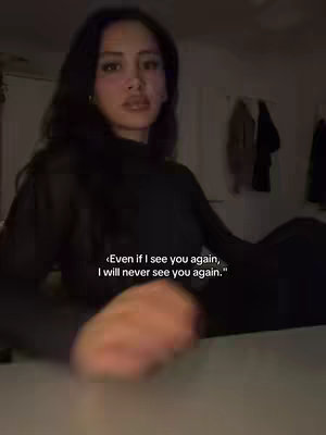 A woman is seated with a dark background, directly facing the camera. She has long, dark hair and is wearing a dark outfit. Text overlayed on the image conveys a somber message.